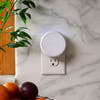 Smart home device attached to a wall socket on a marble surface with fruits and a plant in the foreground.