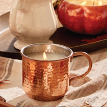 Candlelit copper Simmered Cider candle mug and pumpkin-shaped candle on a wooden table with a blurred natural background