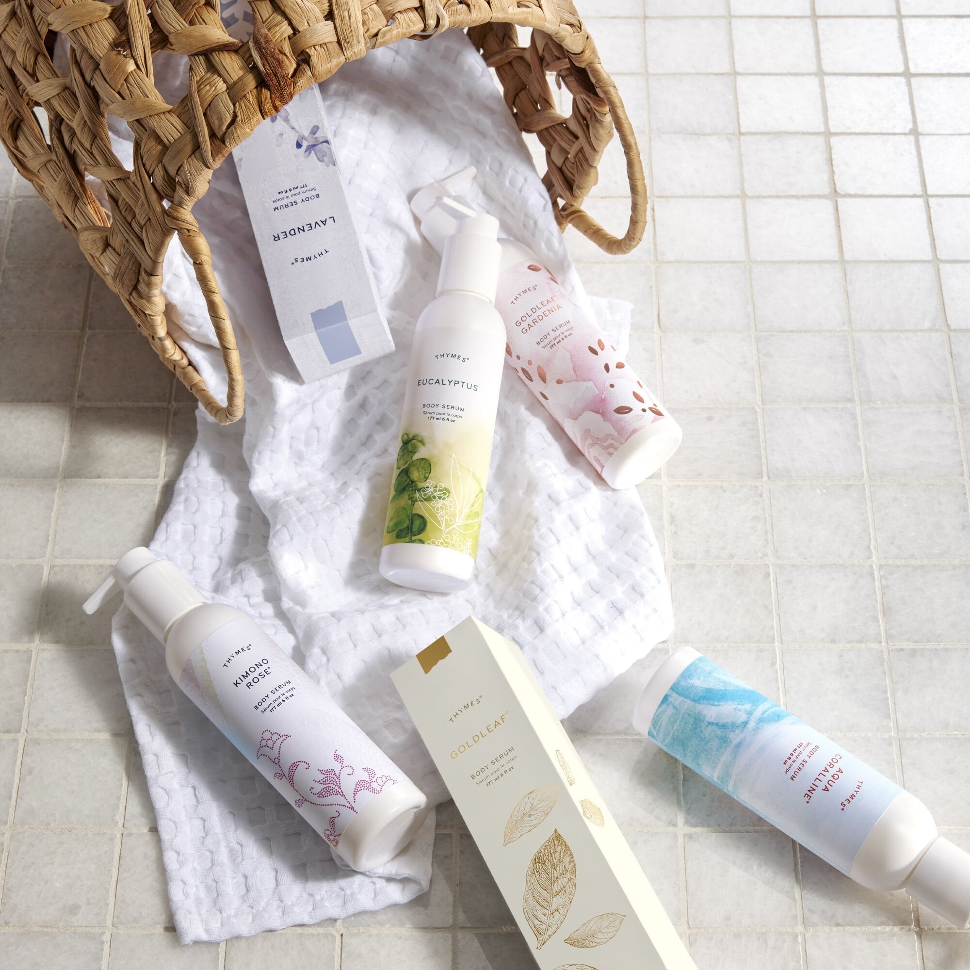 A variety of Thymes Body Serums on top of a towel, coming out of a wicker basket