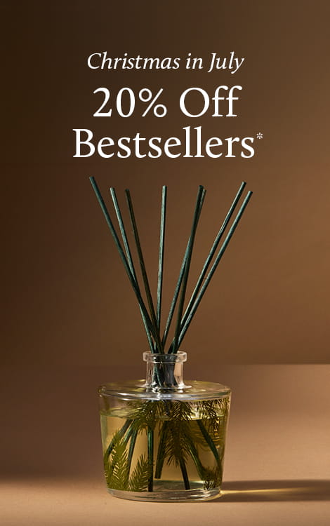 A reed diffuser bottle with dark green reeds and green botanical pieces inside, set against a warm brown background with promotional text reading 'Christmas in July 20% Off Bestsellers'.