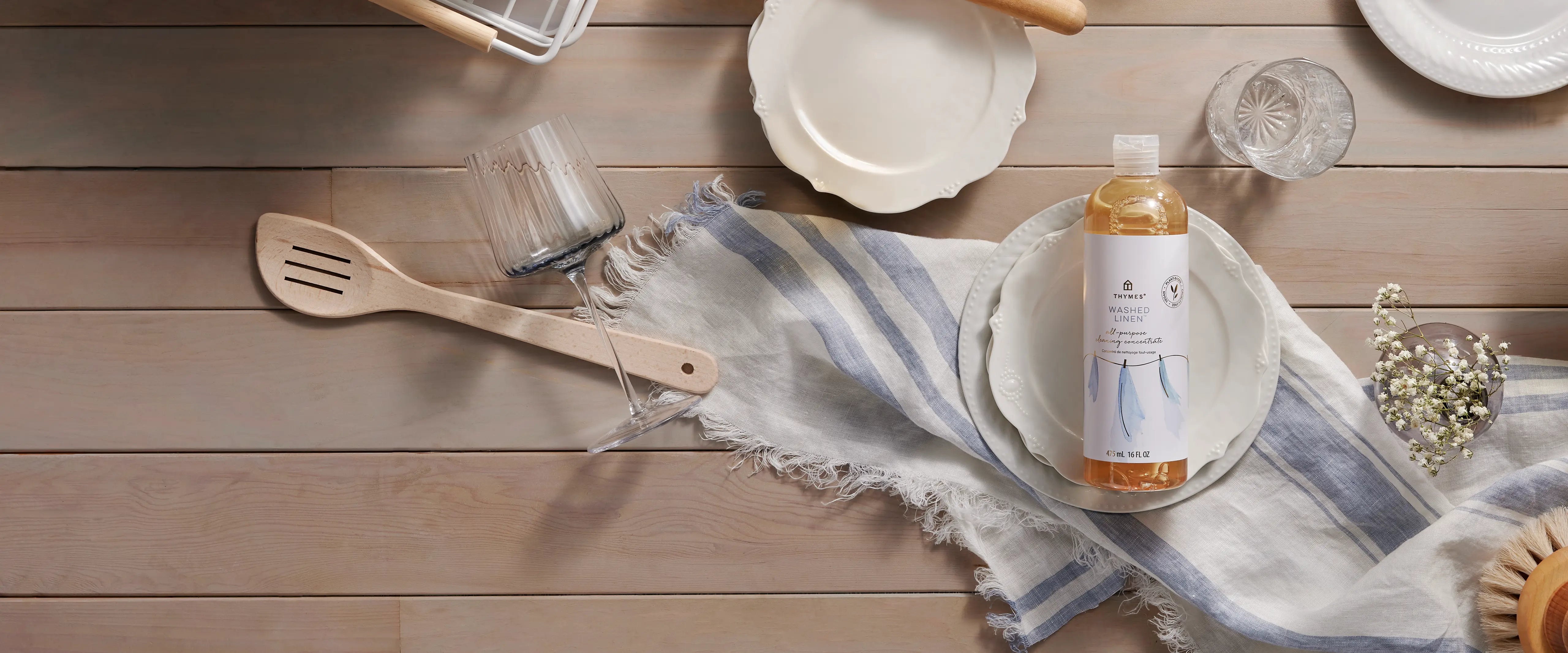 A bottle of dishwashing liquid with a white label and amber liquid inside, styled on a rustic wooden tabletop flat-lay with plates, a wooden spatula, and a striped cloth.