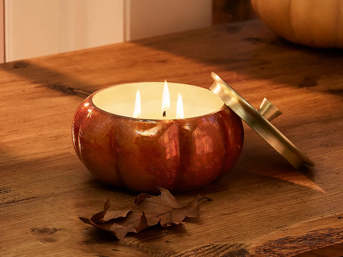 A lit, burnt orange Pumpkin Laurel  Statement Candle on a rustic wooden table with autumn leaves.