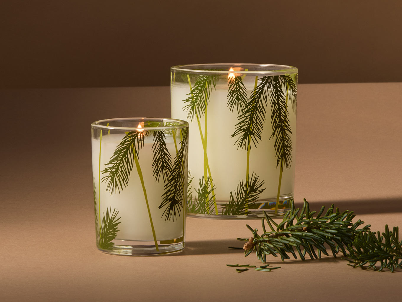 Two glass candles with greenery inside on a brown background