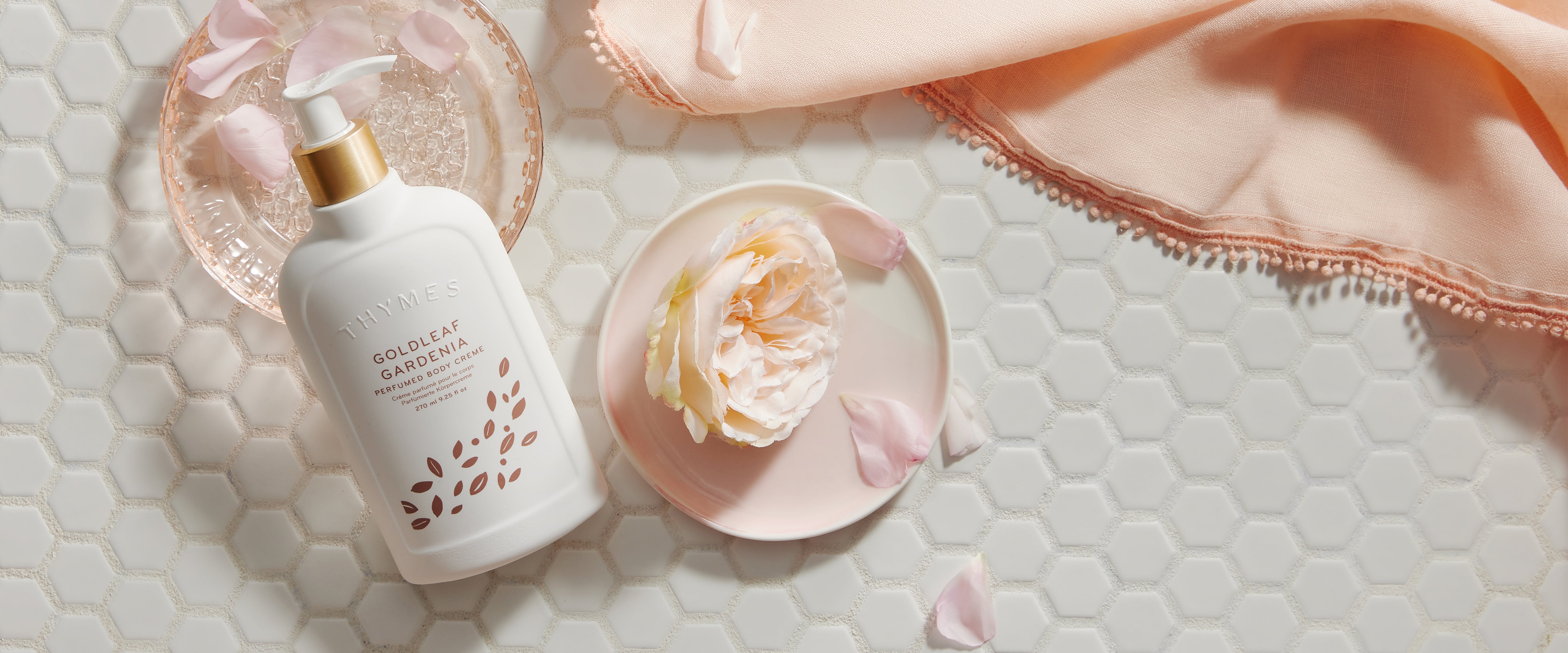 White bottle with gold cap on a hexagonal tile surface with a pink flower and petals.