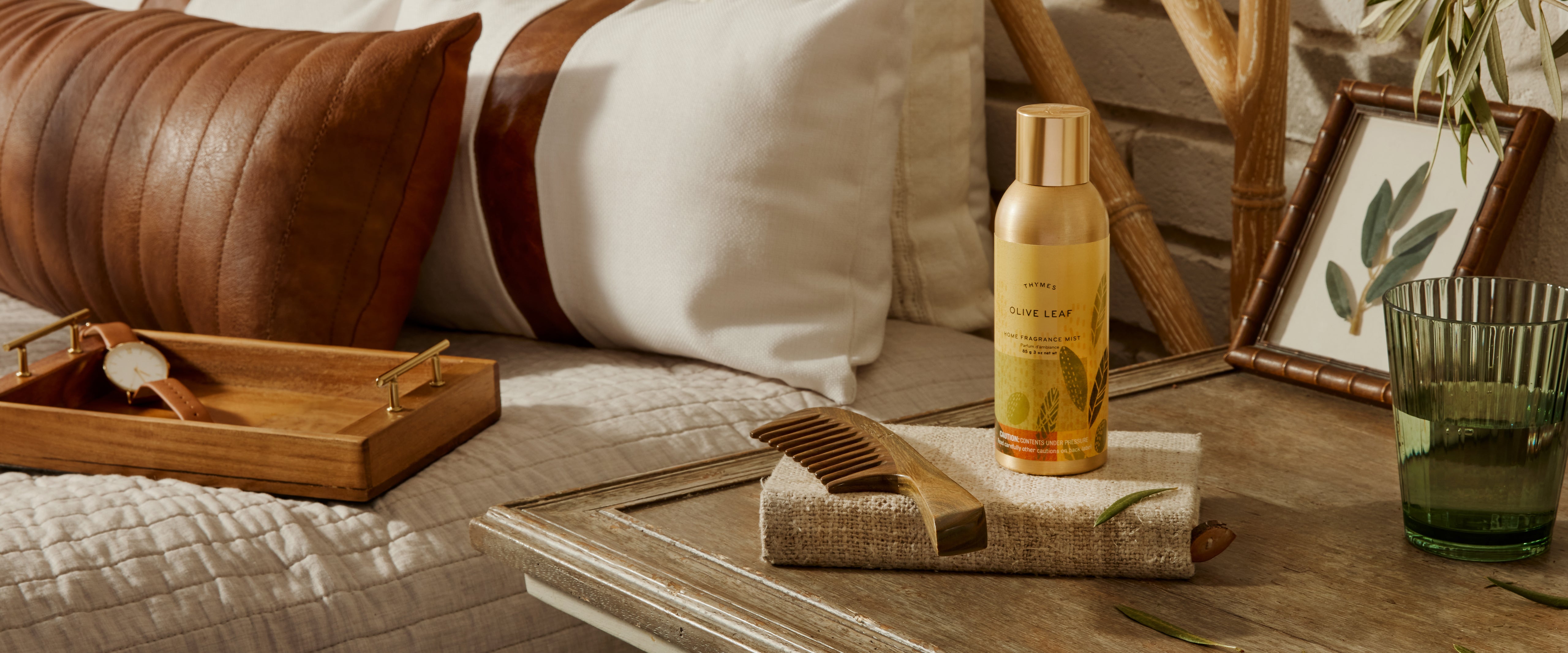 Bottle of body wash on a tray with a comb and brush on a wooden surface, surrounded by pillows and decor.