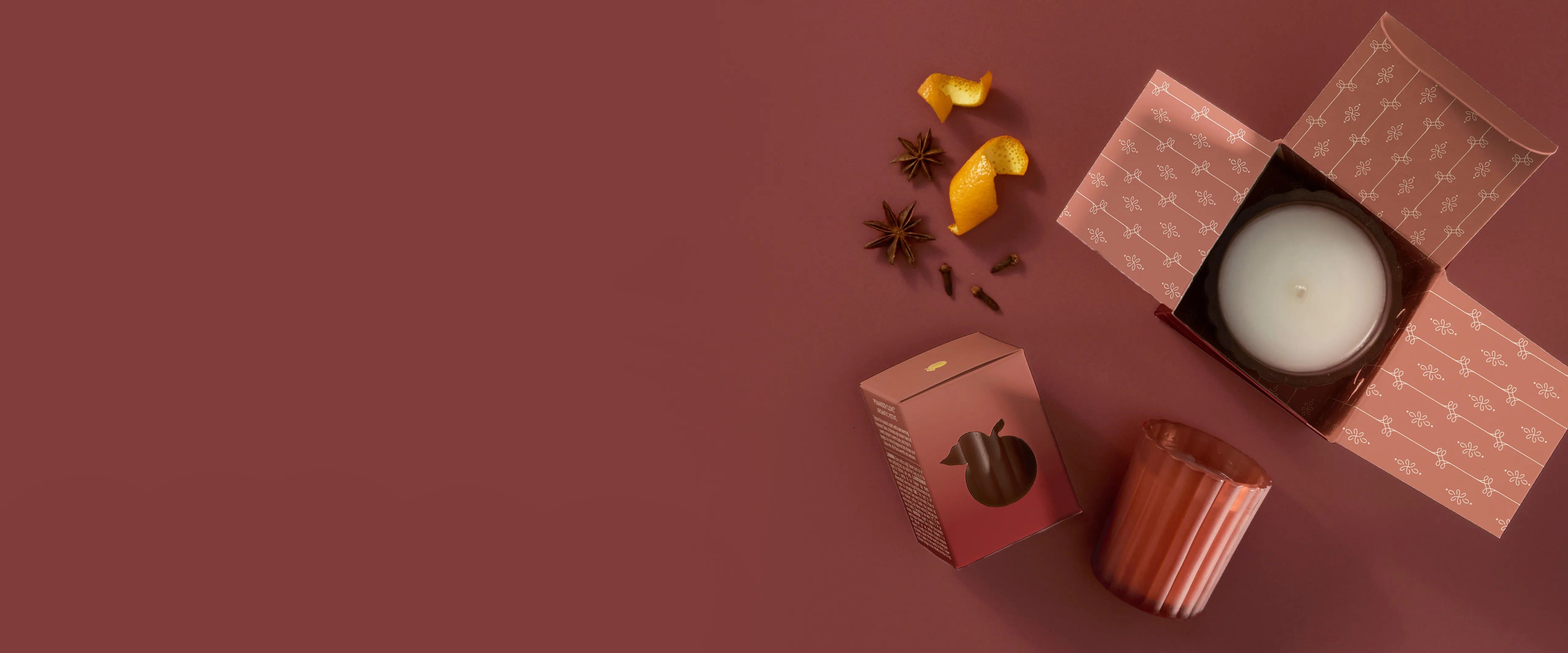 A 6.1 ounce Poured Candle in  an open box, two Votive Candles: one boxed, one unboxed. Mandarin orange peel, star anise, and whole cloves are beside the candles.