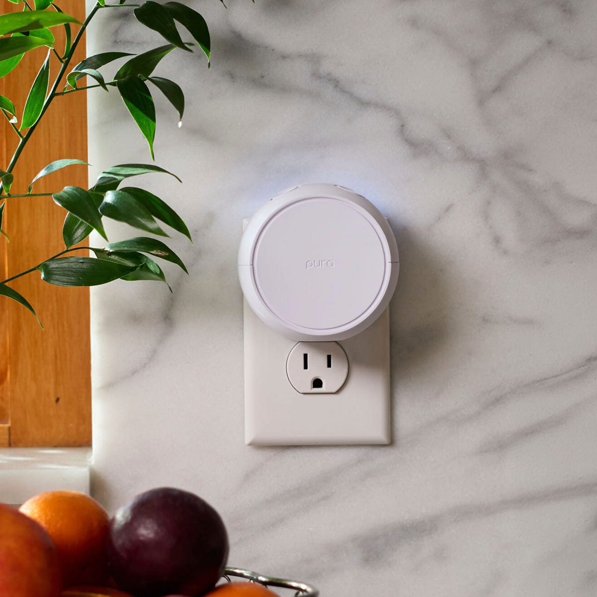 Alternate image of Smart home device attached to a wall socket on a marble surface with fruits and a plant in the foreground.