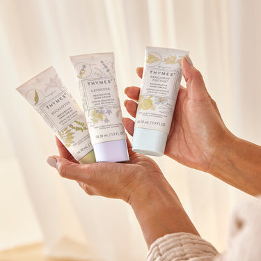 Three bottles of Thymes hand cream held by a person against a neutral background