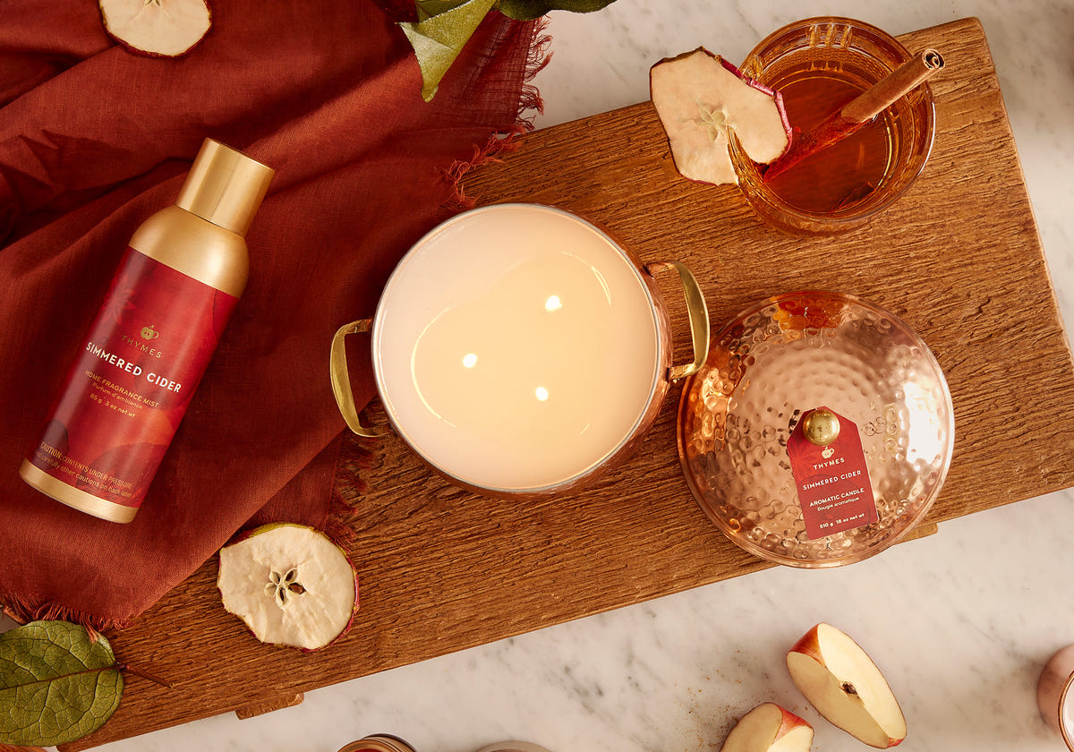 Variety of simmered cider home fragrance products on a plank of wood surrounded by sliced apples and apple cider glass