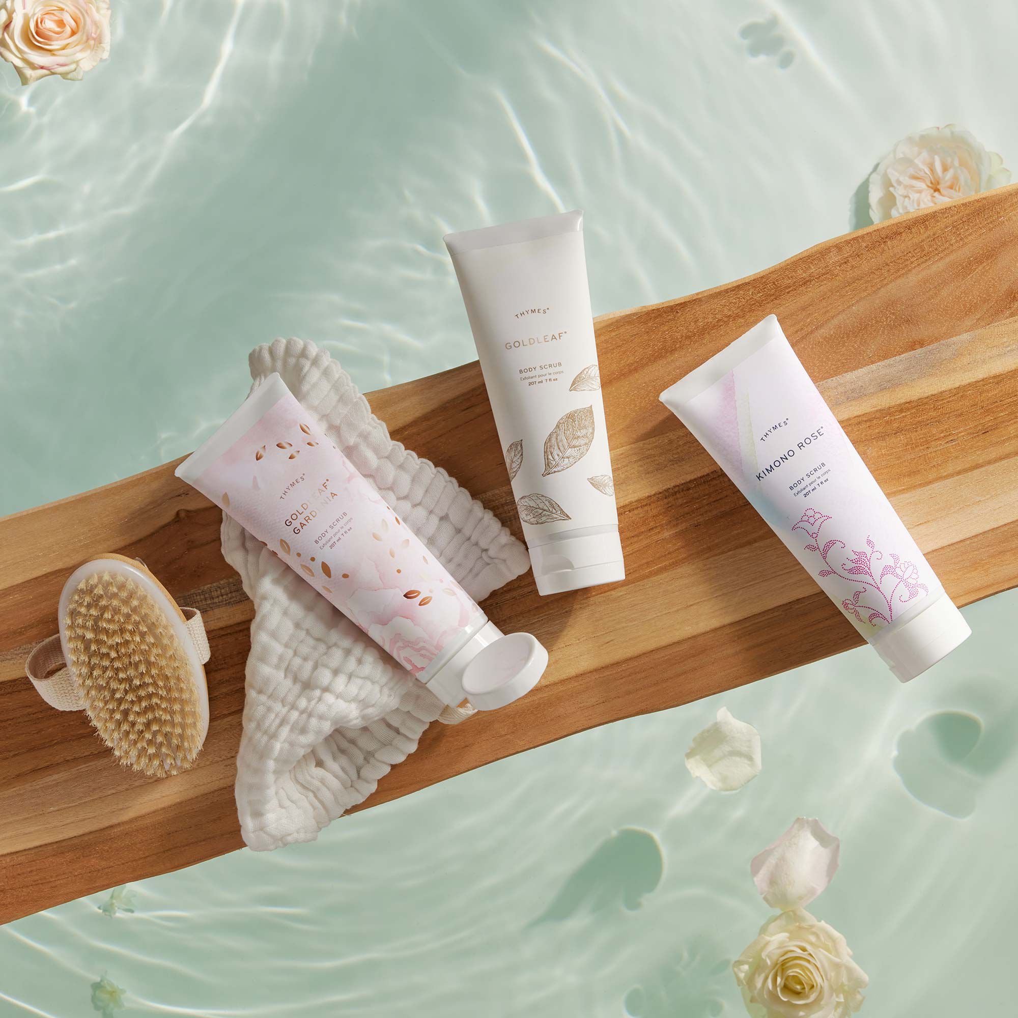 Thymes Kimono Rose Body Scrub for exfoliation laying next to other Thymes Body Scrubs on wooden bath tray in bath tub