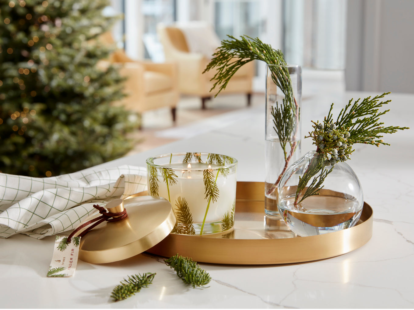 A holiday-themed home decor scene featuring a gold round tray holding a glass jar candle with pine needles inside, a clear glass vase with water and pine branches, and a gold lid resting beside them on a white marble surface.