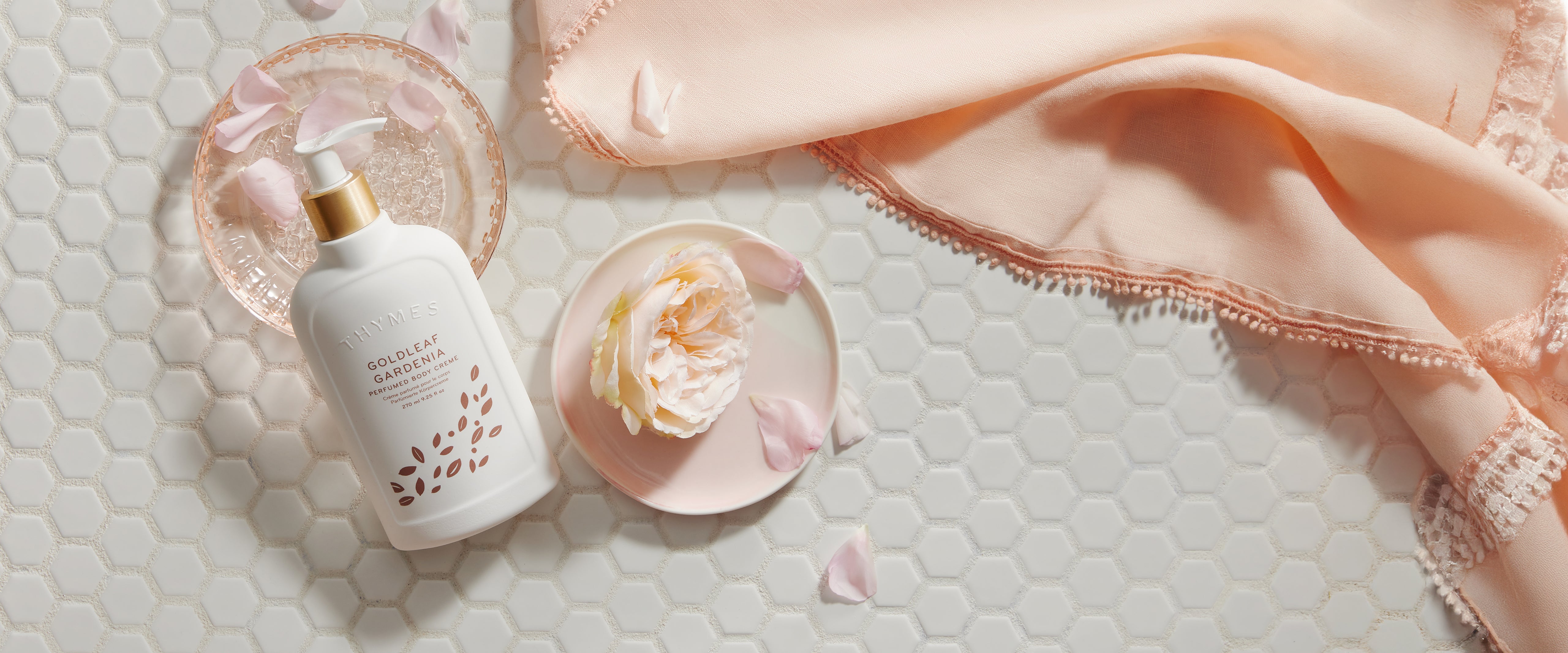 A bottle of Goldleaf Gardenia Body Cream on a tiled surface surrounded by flower petals