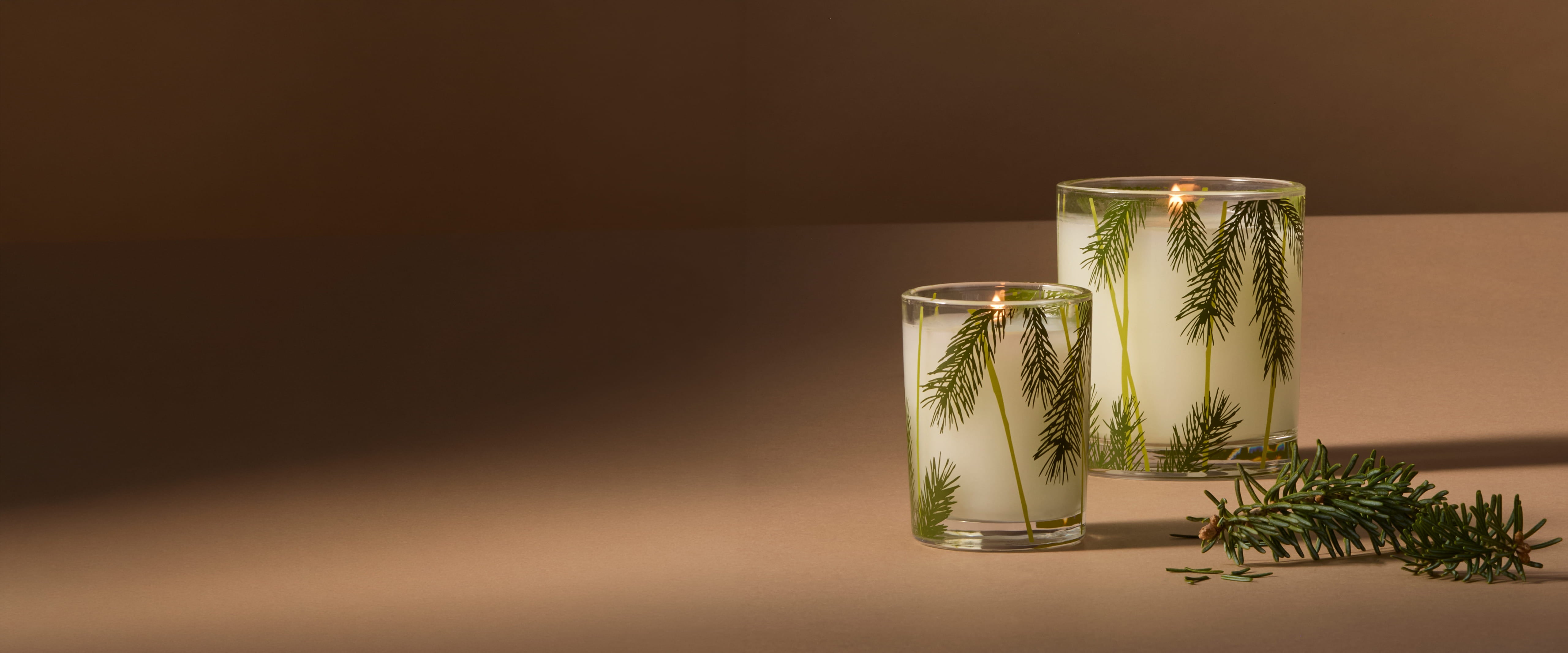 Two clear glass jar candles with white wax and green botanical sprigs inside, lit and set on a warm brown surface against a matching brown background.