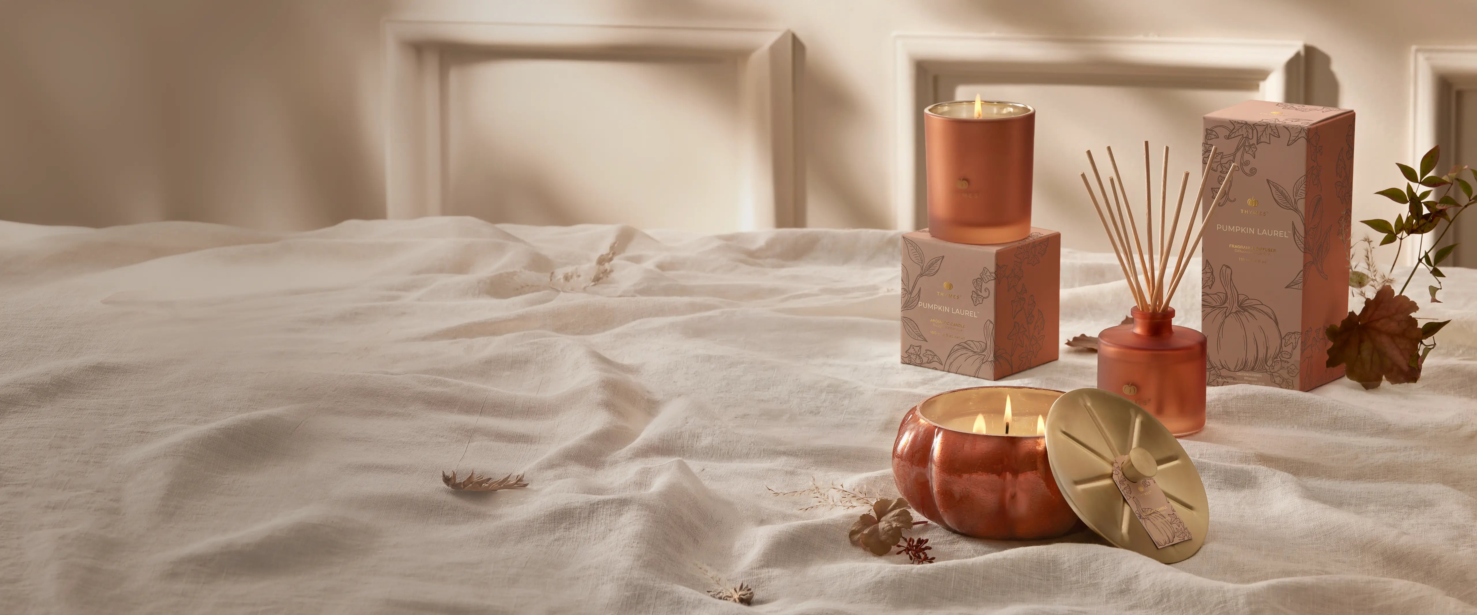 A styled collection of home fragrance products including a lit copper candle, a reed diffuser with reeds, and a matching diffuser box, arranged on a rumpled white bedspread with a neutral wooden headboard in the background.