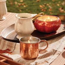 Candlelit copper Simmered Cider candle mug and pumpkin-shaped candle on a wooden table with a blurred natural background