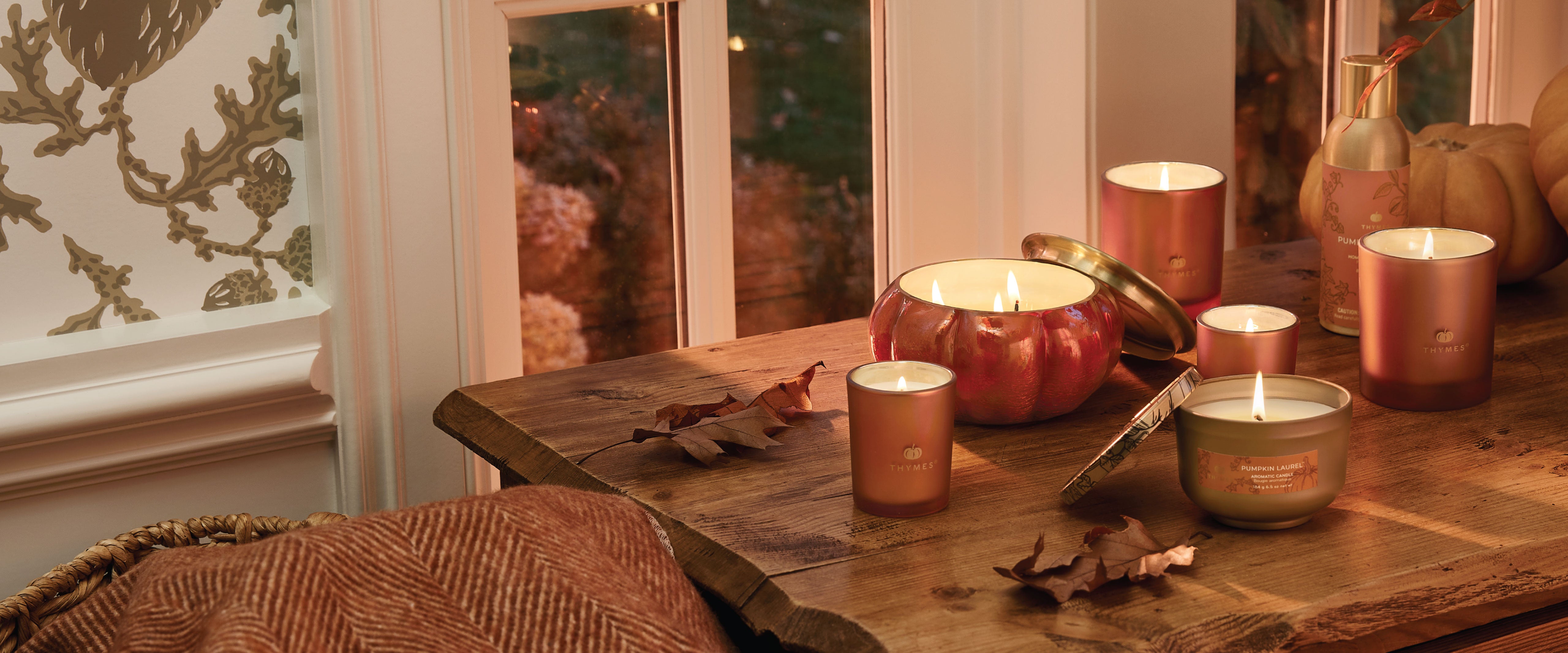 Thymes candles in pumpkin-shaped holders on a wooden table with a warm, cozy atmosphere.