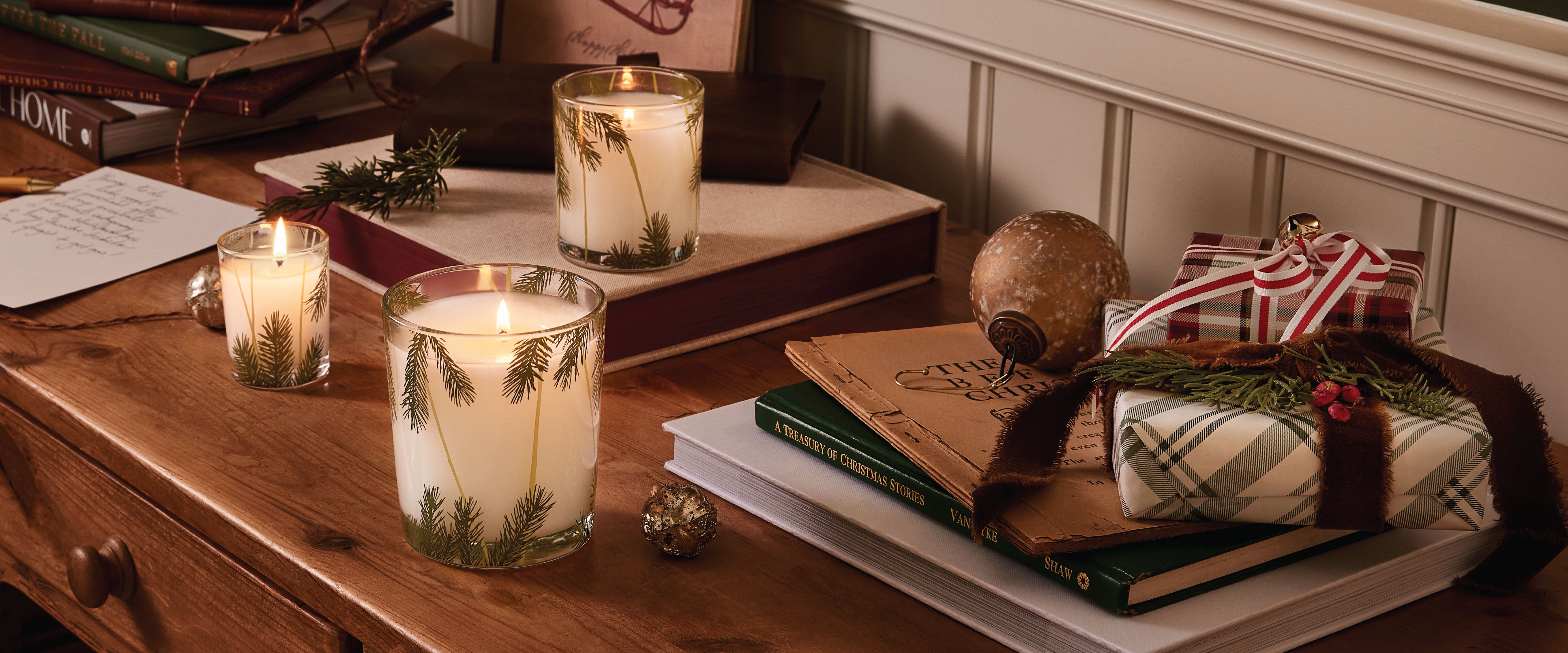 Three candles with pine needle designs on a wooden surface with books and decorative items.