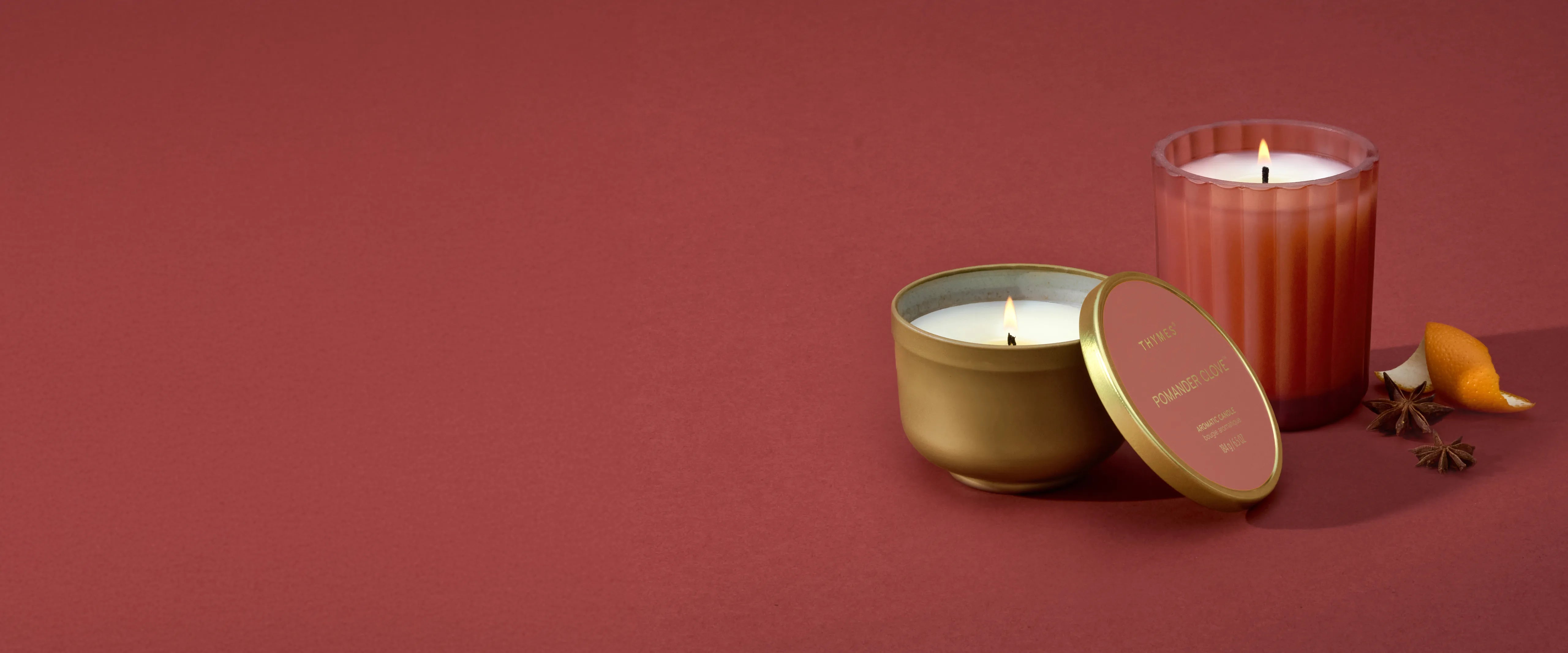 A lit and open Pomander Clove Candle Tin with the lid resting against it and a lit Pomander Clove Poured Candle beside star anise pods and mandarin peel.