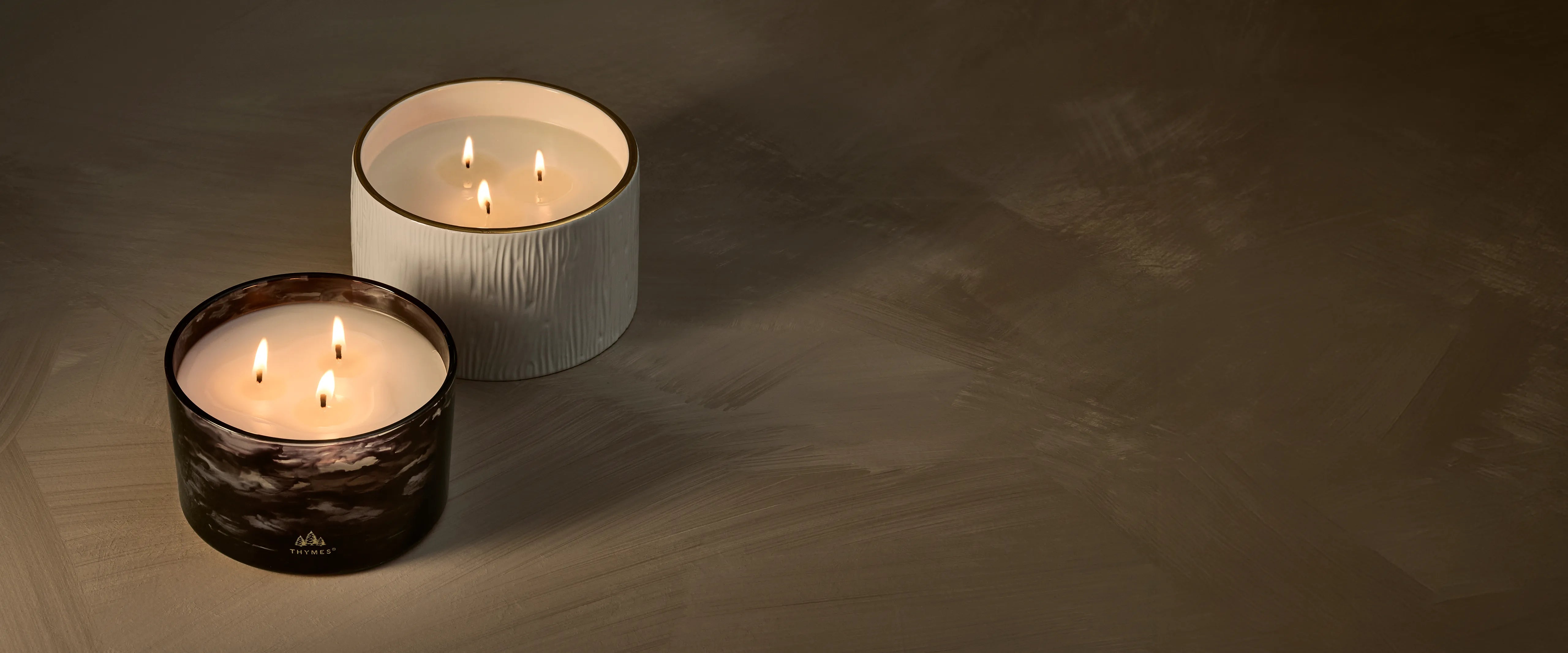 A lit, Noir Woods 3-Wick Candle in a marbled, smoky black and grey vessel beside a lit, Frasier Fir Ceramic 3-Wick Candle in a textured, white ceramic vessel with a 24-karat gold rim.