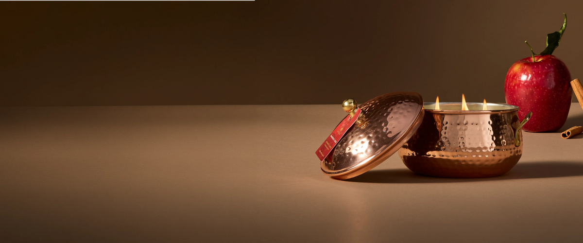 A lit, 3-wick Simmered Cider candle in a copper pot with gold accents, Its lid resting against it.  Displayed with a whole, red apple and cinnamon sticks.