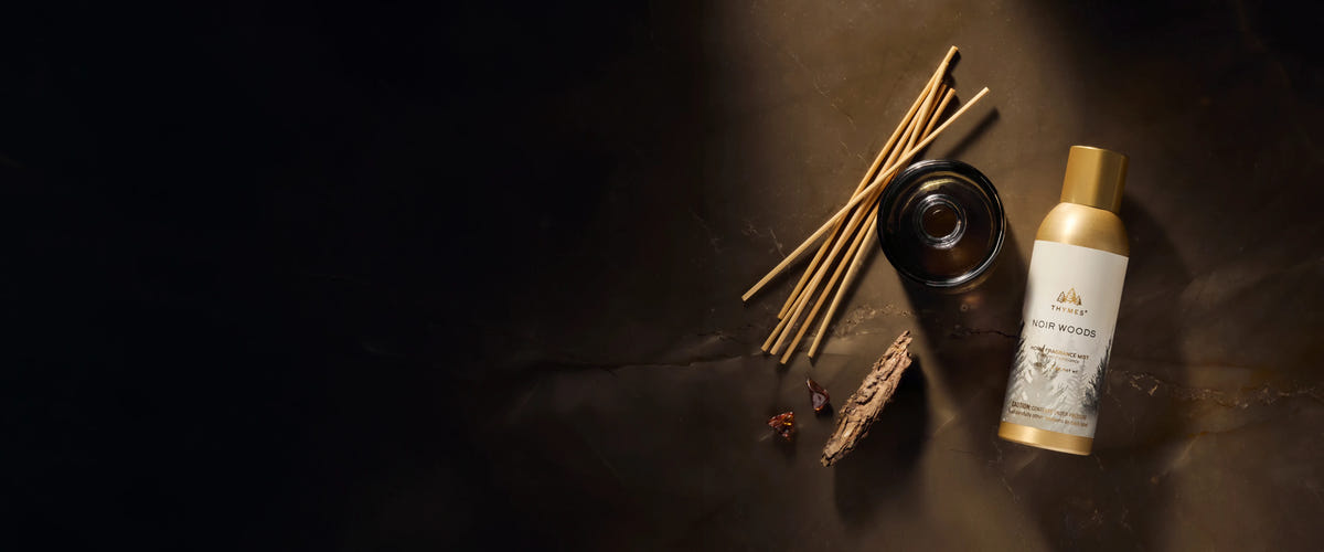 A Noir Woods Reed Diffuser Bottle beside natural wood reeds, a Noir Woods refill bottle, amber shards, and tree bark.