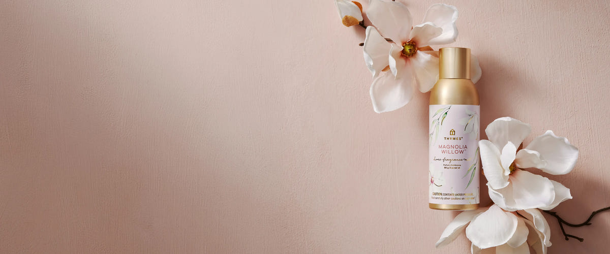 Thymes Magnolia Willow room spray in a gold bottle surrounded by white magnolia flowers against a blush pink background.
