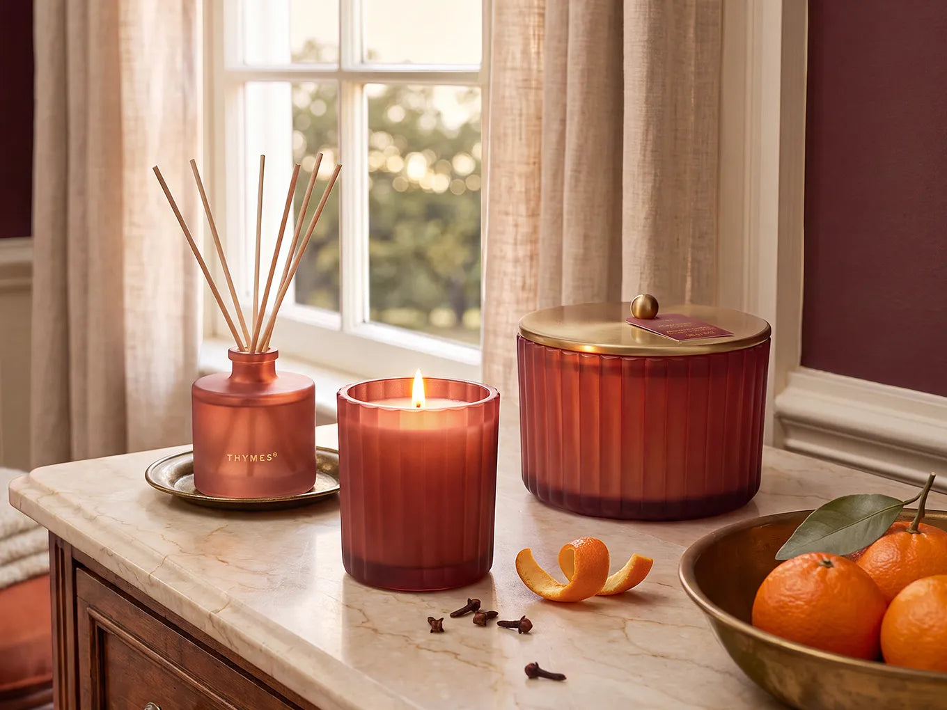 A reed diffuser, a lit 6.1 ounce candle and a covered 3 wick candle on a marble-topped cabinet with cloves, mandarin peels and a bowls of mandarin oranges.