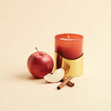 Thymes Simmered Cider Harvest Red Poured Candle with Gold Sleeve next to apple and cinnamon stick