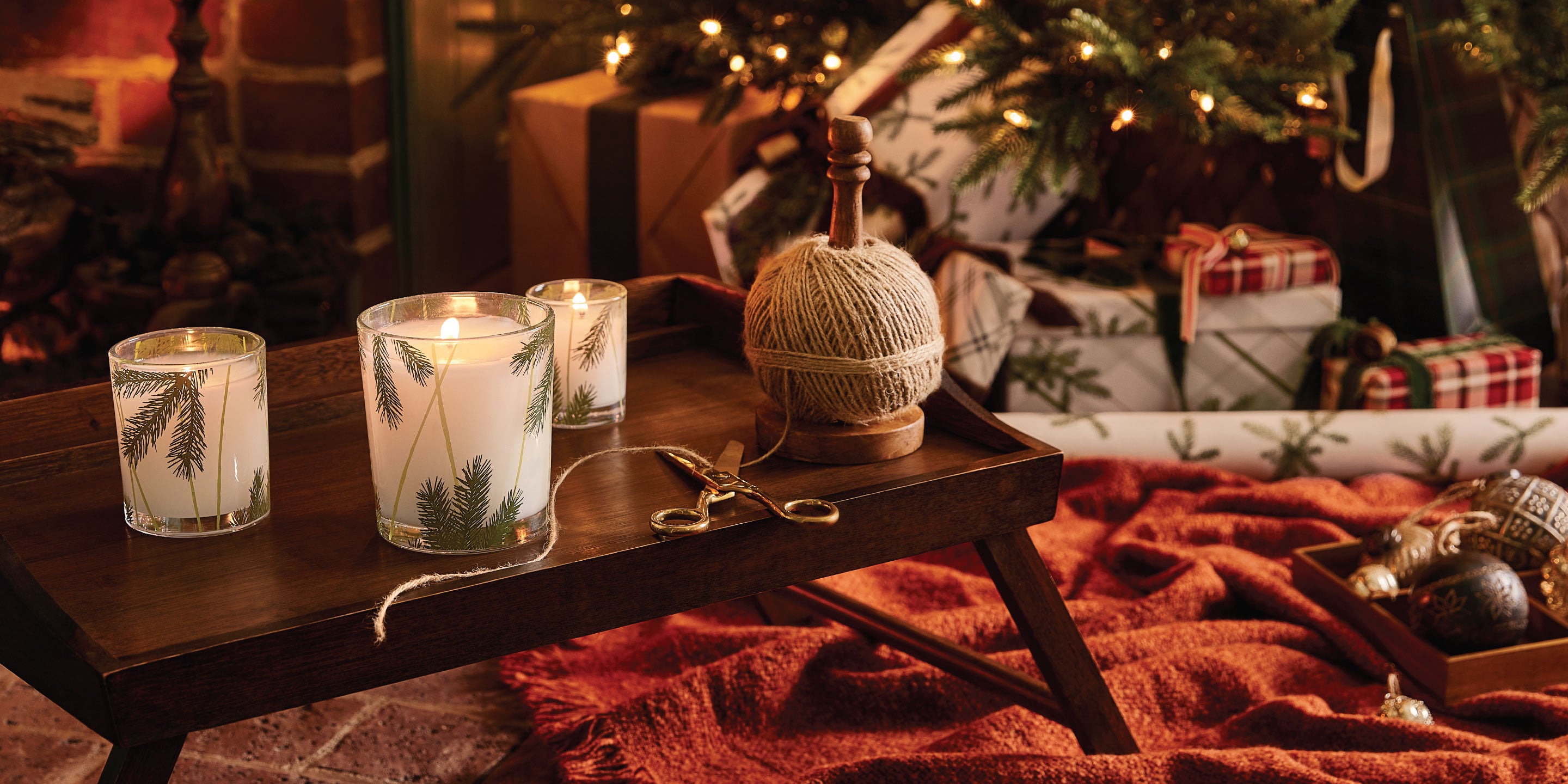 Candles on a wooden table with a cozy Christmas setting featuring a tree and presents.