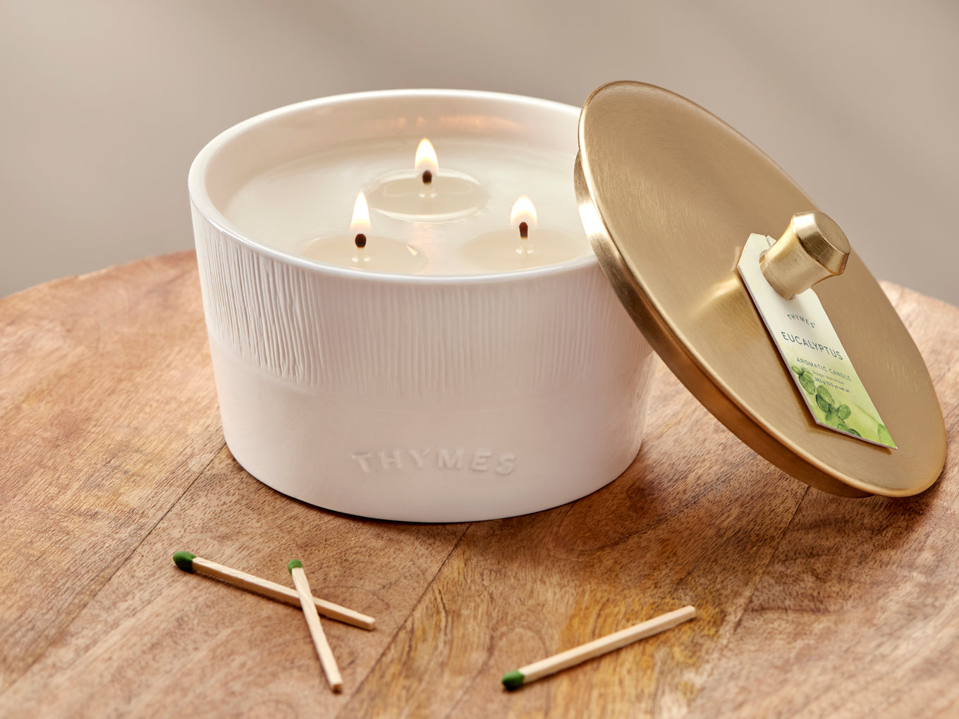 White candle with gold lid on a wooden surface, surrounded by matches.