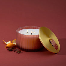 A  lit 3 Wick candle in a frosted, matte, sienna orange vessel with its metallic, gold finish lid resting against it.  Mandarin peel, star anise, and whole cloves are beside it.