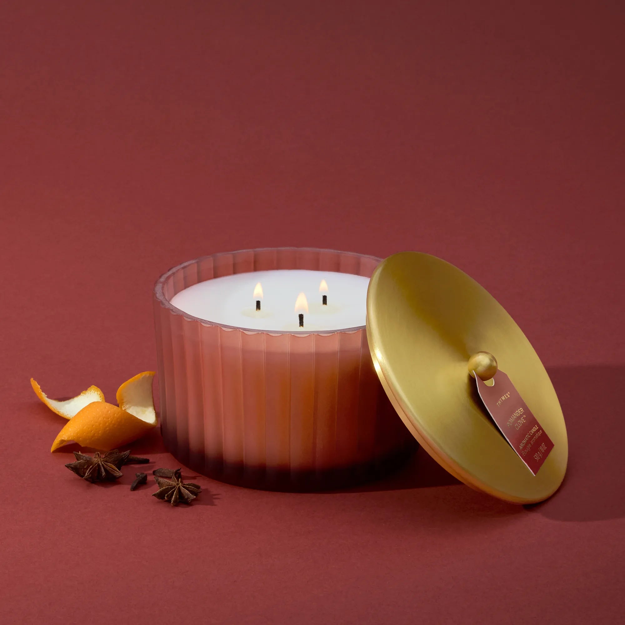 A  lit 3 Wick candle in a frosted, matte, sienna orange vessel with its metallic, gold finish lid resting against it.  Mandarin peel, star anise, and whole cloves are beside it.
