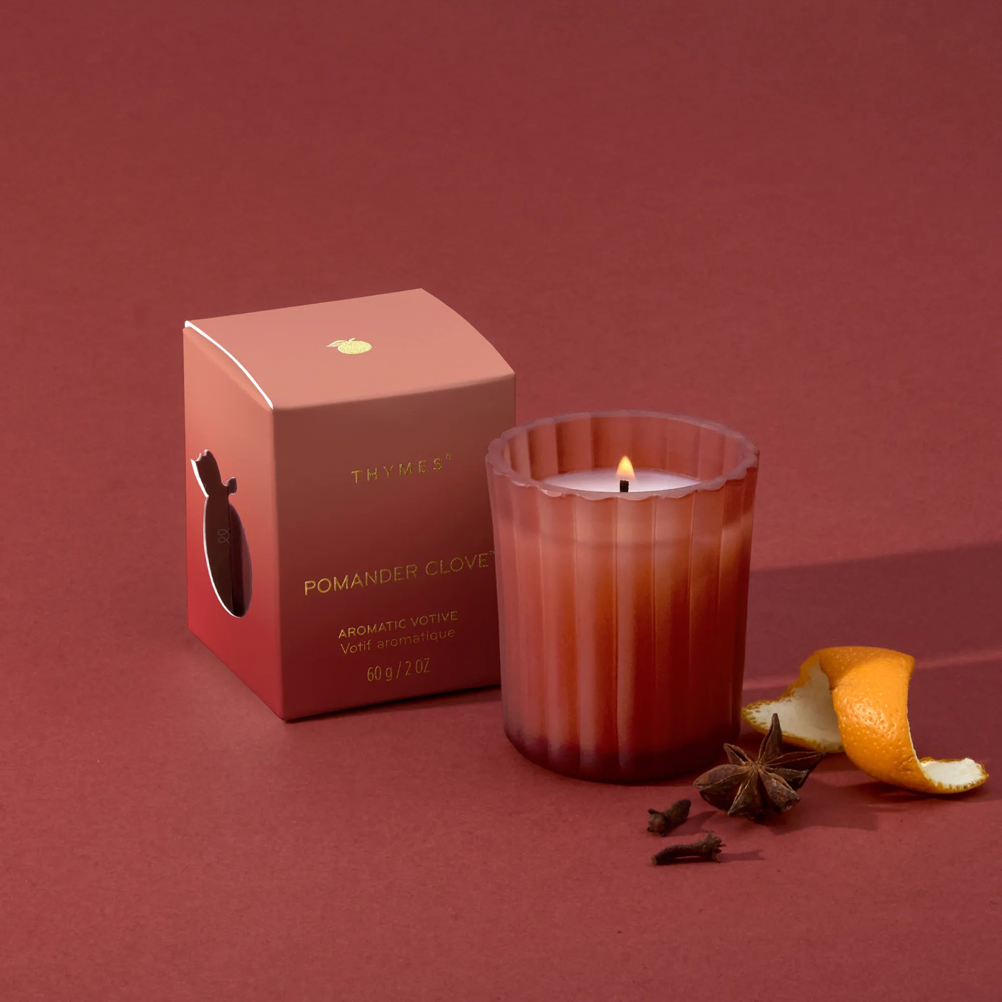 A lit, 2 ounce votive candle in a frosted, matte glass vessel beside its matching packaging, mandarin peel, star anise, and whole cloves.