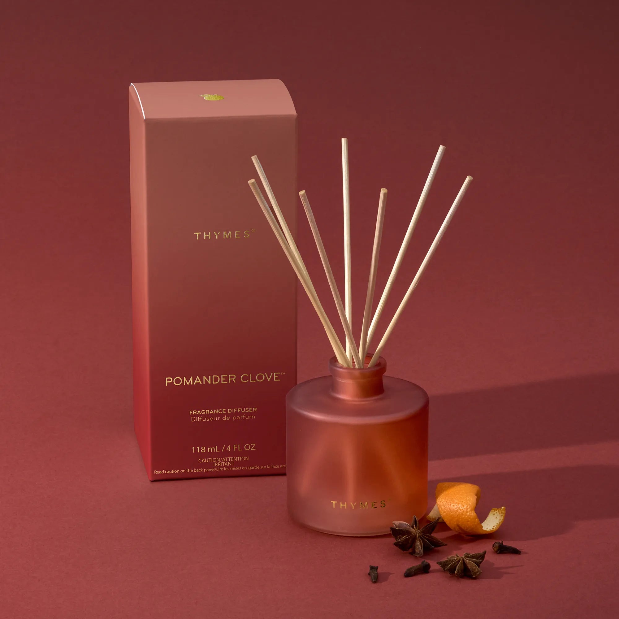 A frosted, matte, sienna orange glass reed diffuser bottle with inserted reeds beside its packaging, a mandarin peel, star anise, and whole cloves.