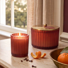 A lit, poured candle in a frosted, matte, sienna orange glass vessel beside a matching 3 wick candle with a gold, metallic finished lid, whole cloves, mandarin peel and a bowl of mandarin oranges.