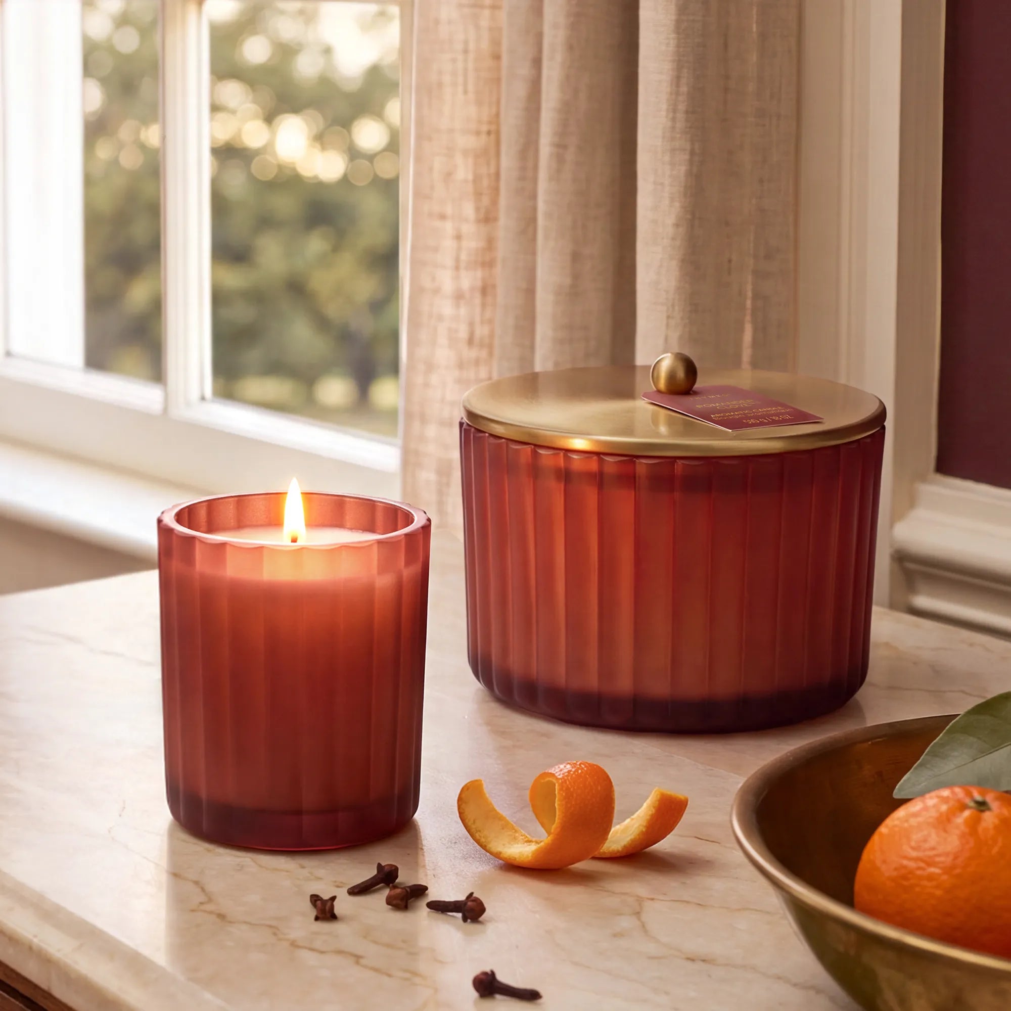 A lit, poured candle in a frosted, matte, sienna orange glass vessel beside a matching 3 wick candle with a gold, metallic finished lid, whole cloves, mandarin peel and a bowl of mandarin oranges.