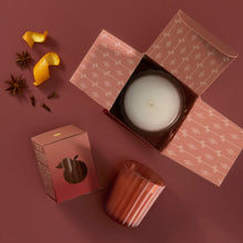 A 6.1 ounce Poured Candle in  an open box, two Votive Candles: one boxed, one unboxed. Mandarin orange peel, star anise, and whole cloves are beside the candles.