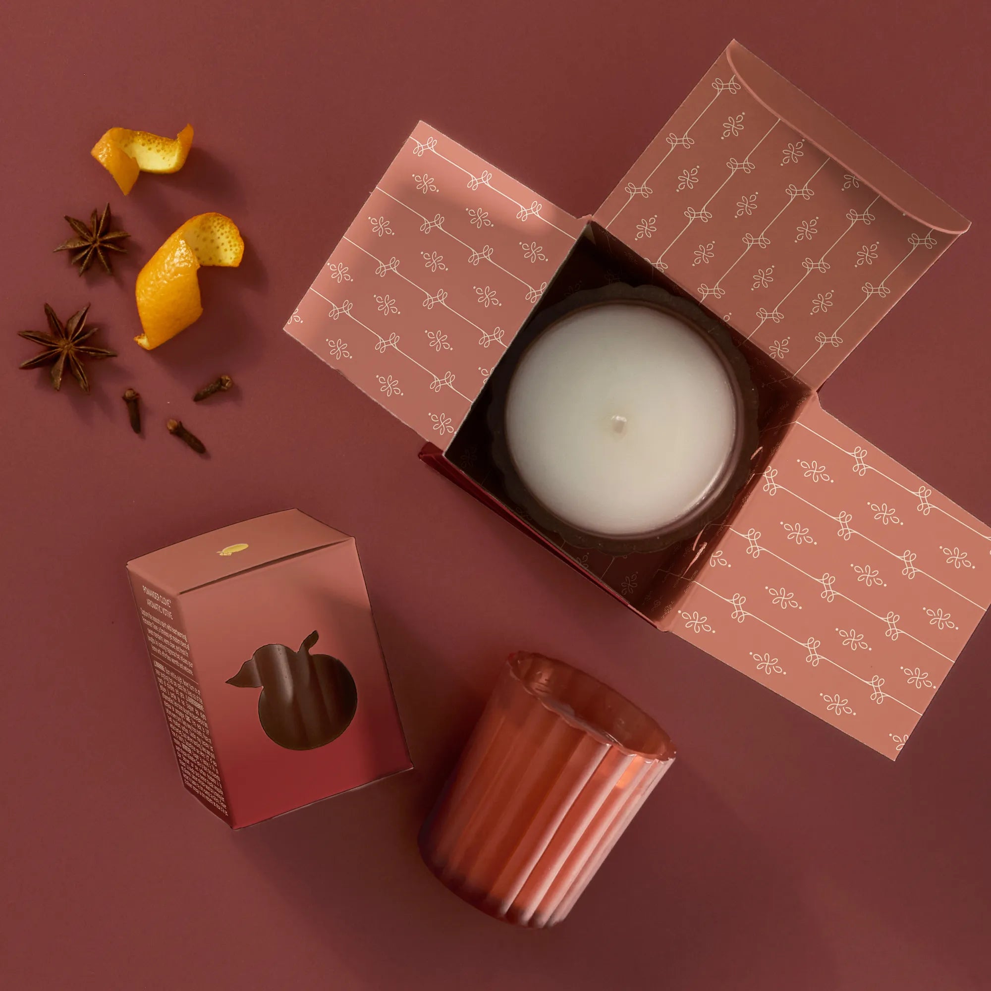 A 6.1 ounce Poured Candle in  an open box, two Votive Candles: one boxed, one unboxed. Mandarin orange peel, star anise, and whole cloves are beside the candles.