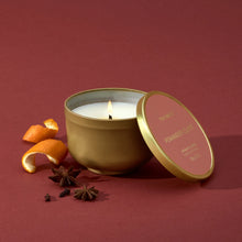 A lit candle in a gold, metallic finish candle tin with its lid resting against it.  Beside it are mandarin peels, star anise and whole cloves.