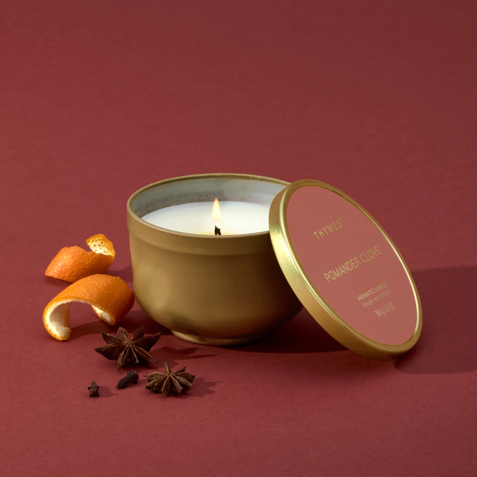 Alternate image of A lit candle in a gold, metallic finish candle tin with its lid resting against it.  Beside it are mandarin peels, star anise and whole cloves.