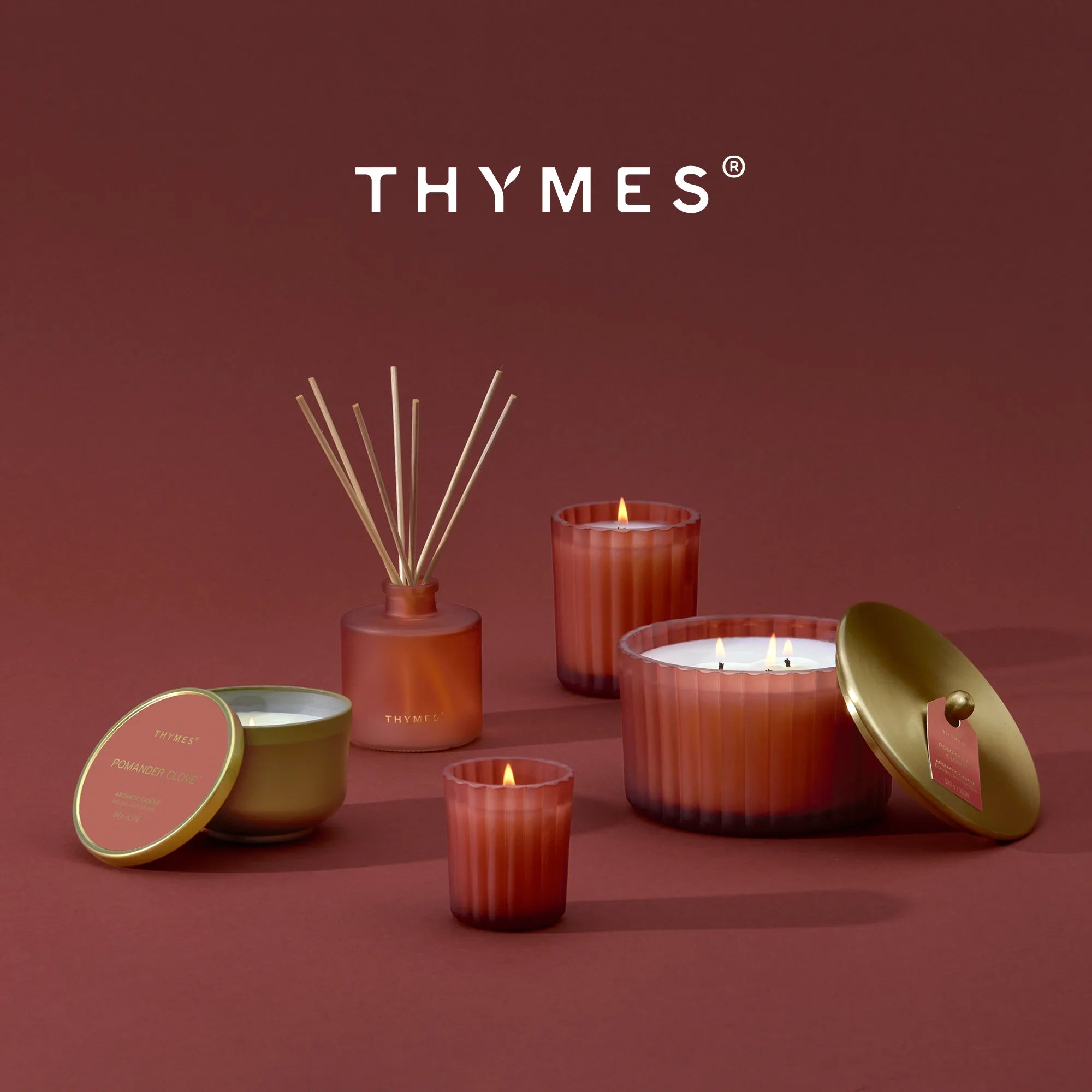 A lit candle in a tin with a gold metallic finish.  A reed diffuser, a lit votive candle, a lit 6.1 oz candle, and a lit 3 wick candle each in frosted, matte, sienna orange vessel.